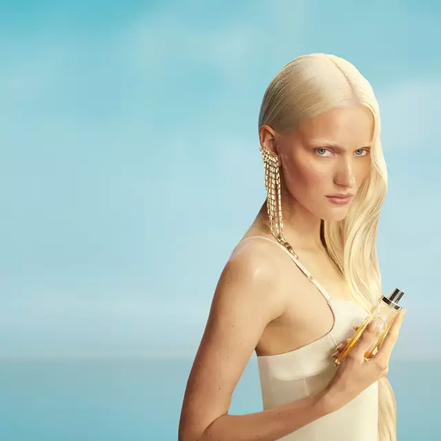 Kampagnenbanner der neuen Mugler Starlicious Les Eaux Gourmandes mit einem Model mit langen blonden Haaren und blauen Augen in einem hellgelben Kleid, das den Pineapple Musk Duftflakon vor einem sanftblauen Himmel hält. Die Düfte sind köstlich, sammelbar und kombinierbar.