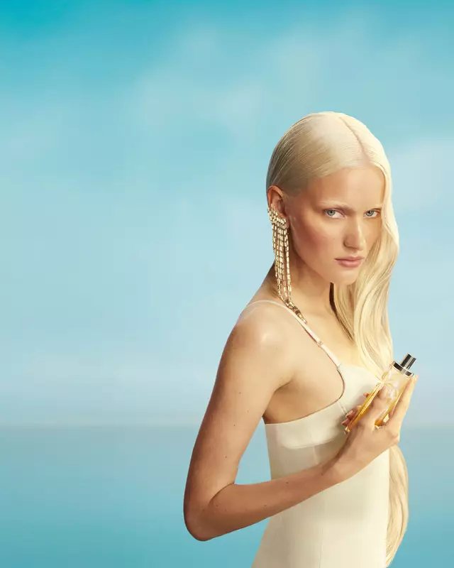 Kampagnenbanner der neuen Mugler Starlicious Les Eaux Gourmandes mit einem Model mit langen blonden Haaren und blauen Augen in einem hellgelben Kleid, das den Pineapple Musk Duftflakon vor einem sanftblauen Himmel hält. Die Düfte sind köstlich, sammelbar und kombinierbar.