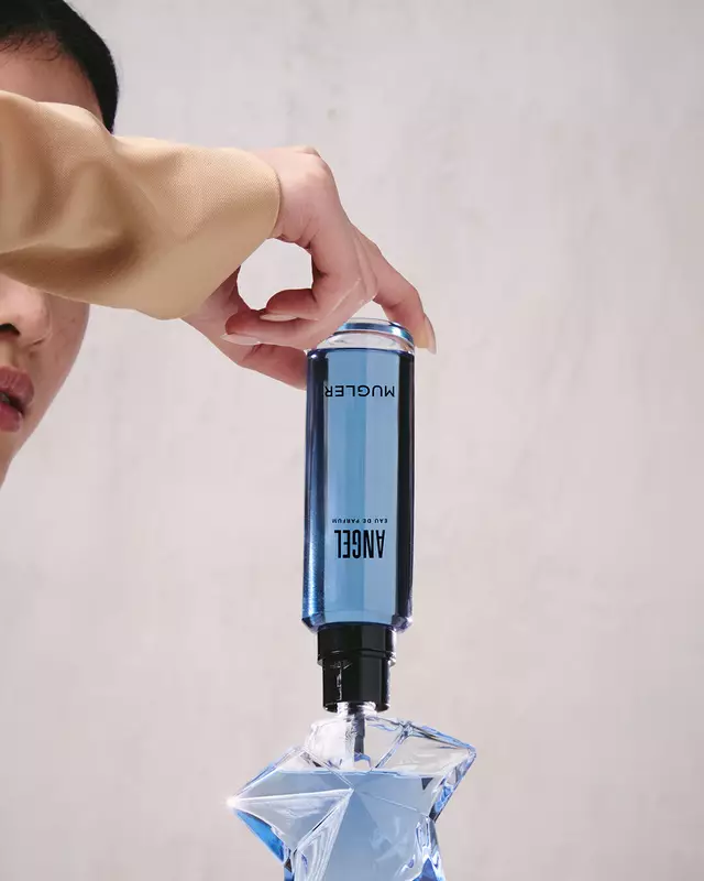 A hand refilling the blue star-shaped Angel Eau de Parfum bottle using a blue refill bottle upside down.